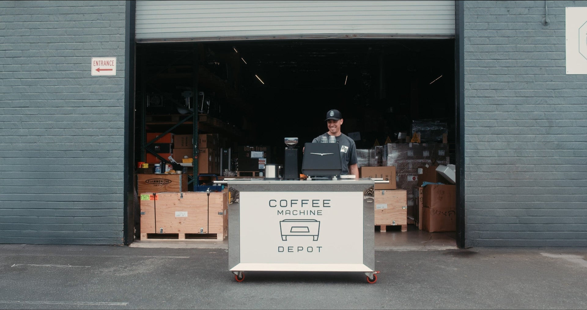 Person standing behind a counter with 'Coffee Machine Depot' branding in front of a warehouse entrance.