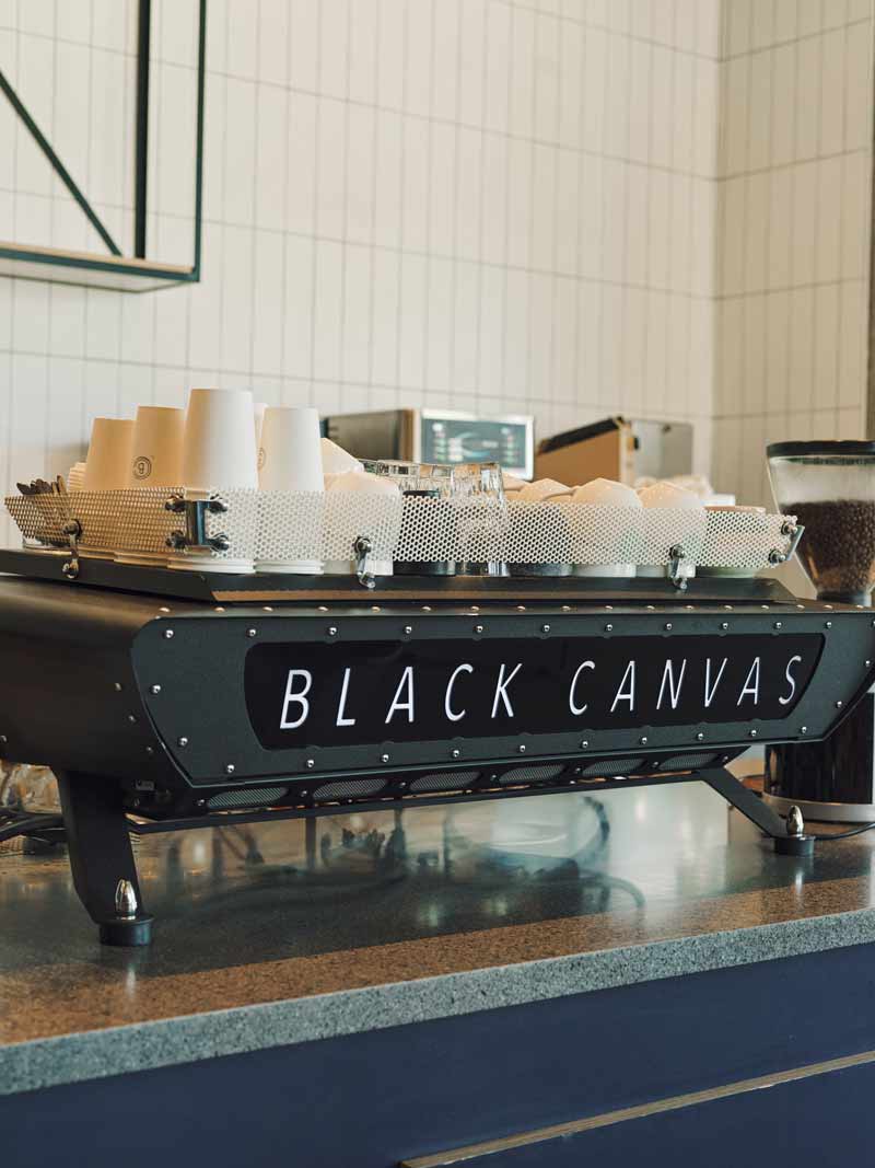 Rear view of a custom Black Canvas espresso machine in a modern café setup, with paper cups and accessories on the cup tray, ready for service