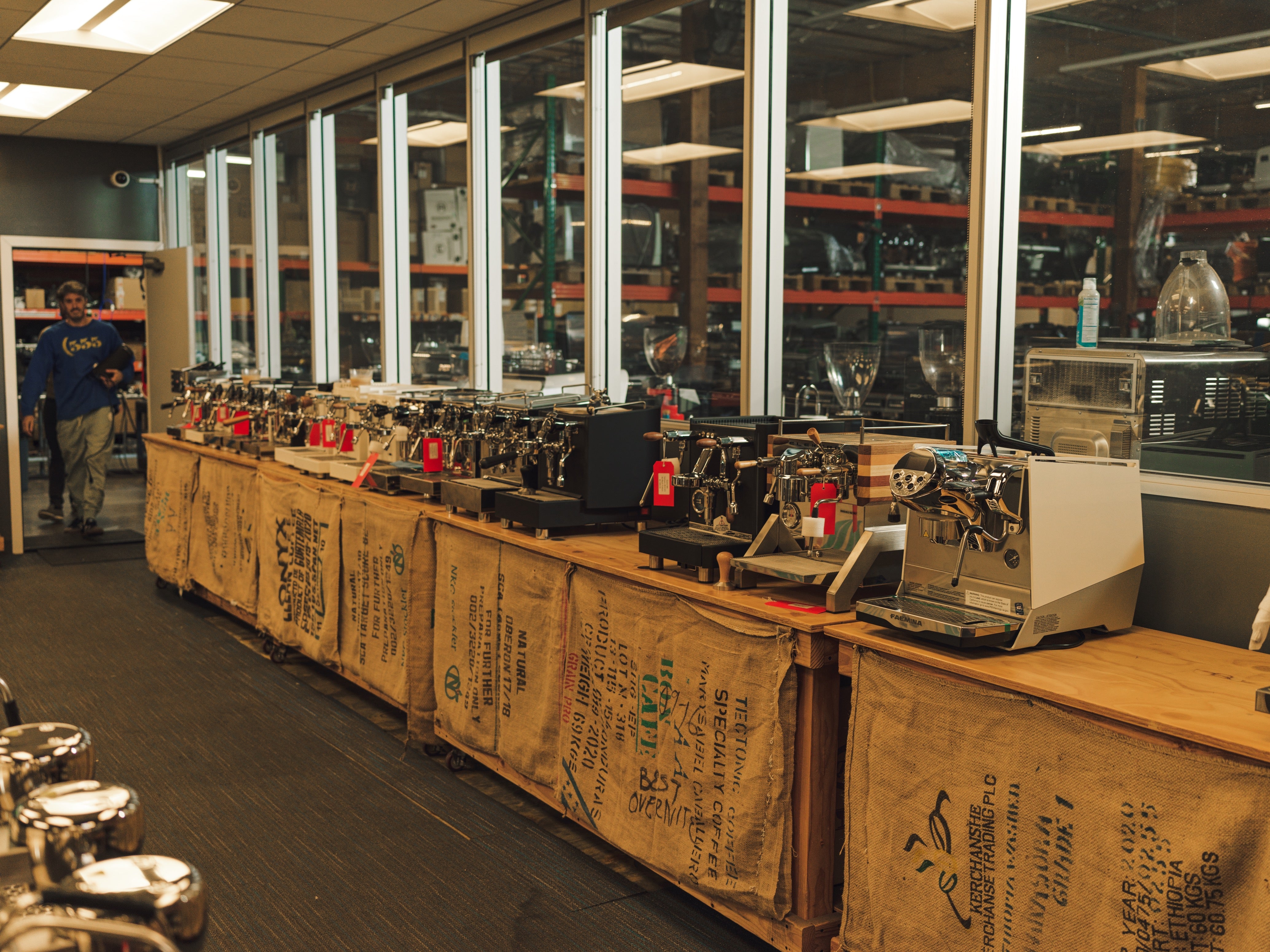 Showroom display of various home and commercial espresso machines on reclaimed wood counters covered with burlap coffee sacks, inside a specialty coffee equipment warehouse.