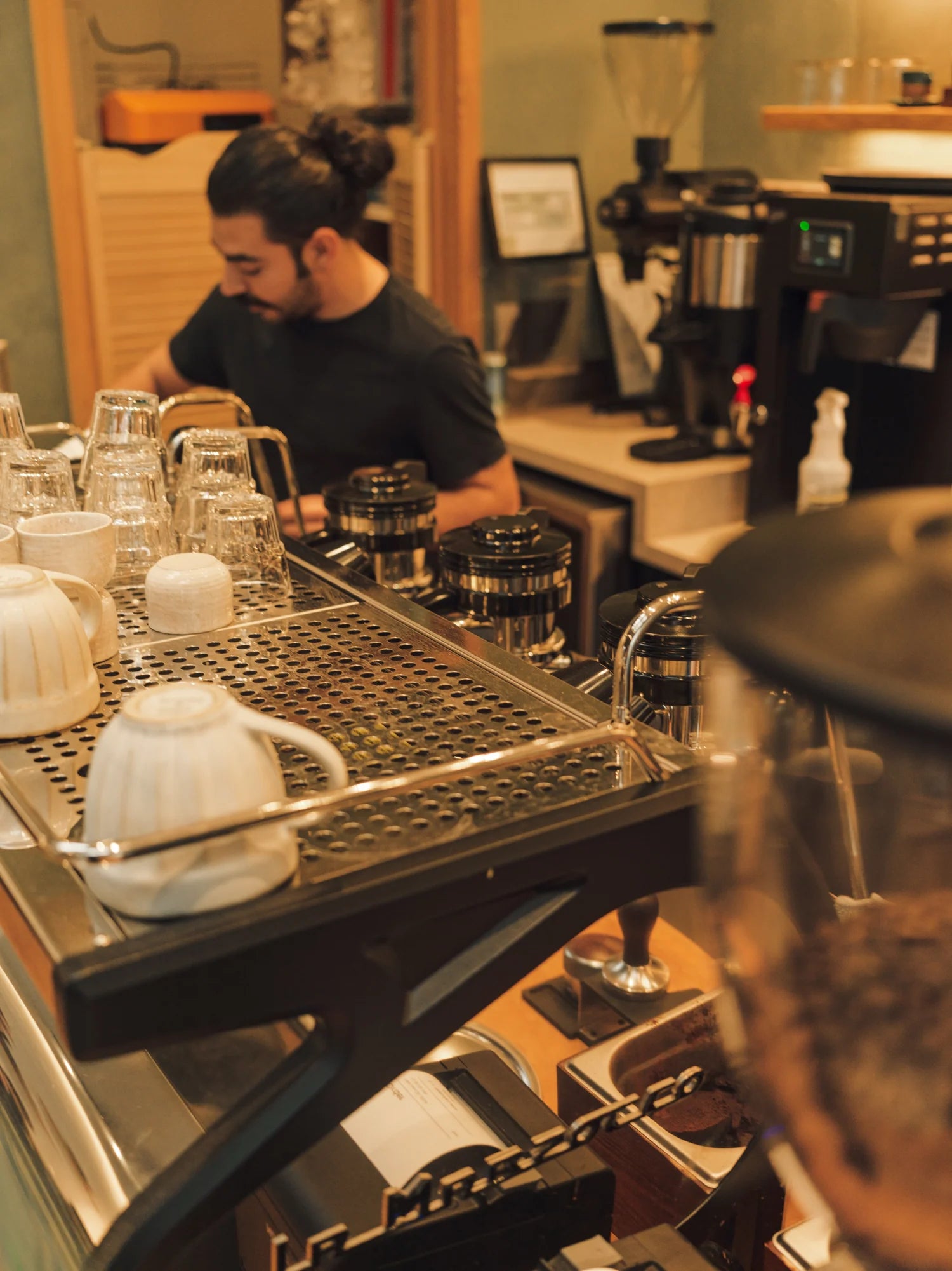 An silver espresso machine with coffee mugs on it, there is also a guy with long hair in the background using the espresso machine.