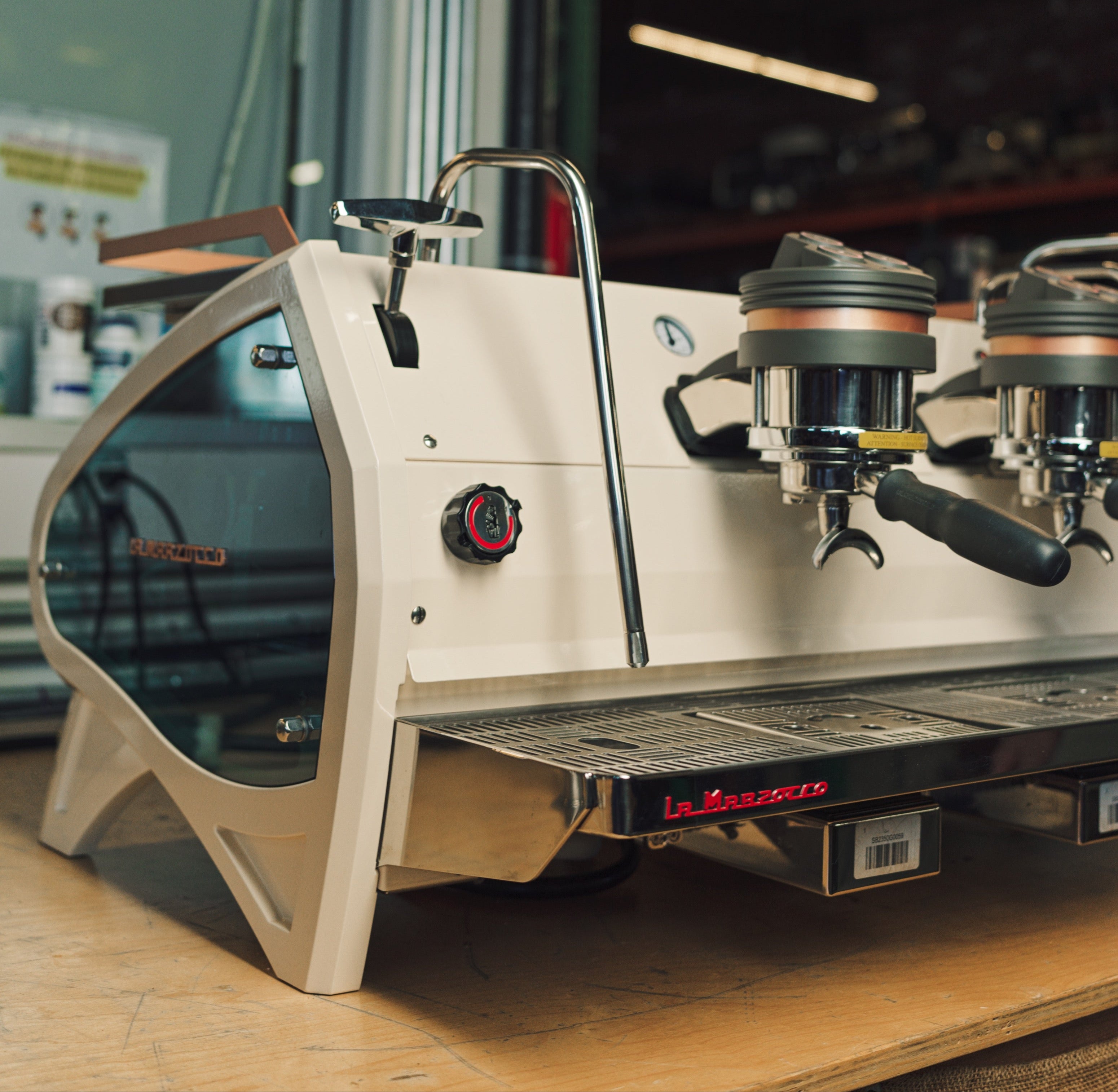 A close-up of a white La Marzocco espresso machine on a wooden countertop, featuring dual portafilters and a chrome steam wand, with a modern industrial background.