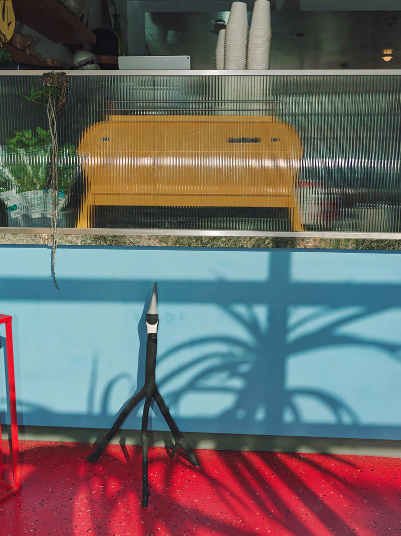 Yellow La Marzocco espresso machine behind textured glass in a colorful café interior with red terrazzo flooring, blue wall, and modern plant decor