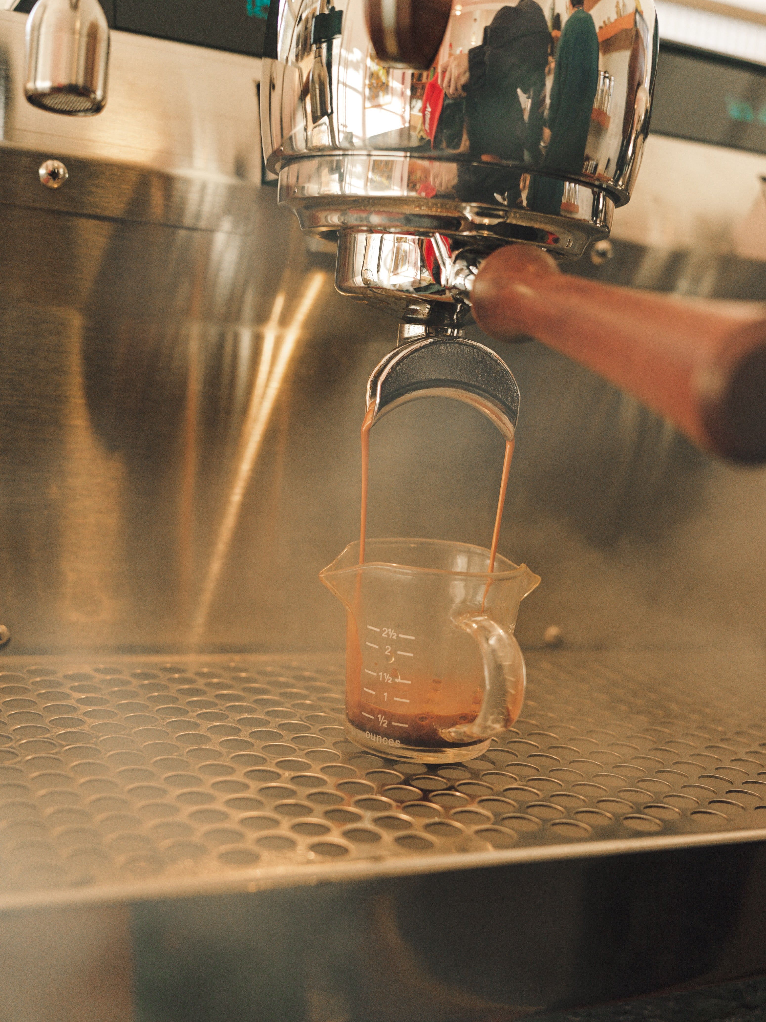 Espresso shot being pulled from a commercial portafilter into a glass measuring cup on a stainless steel drip tray, surrounded by steam in a professional café setting.